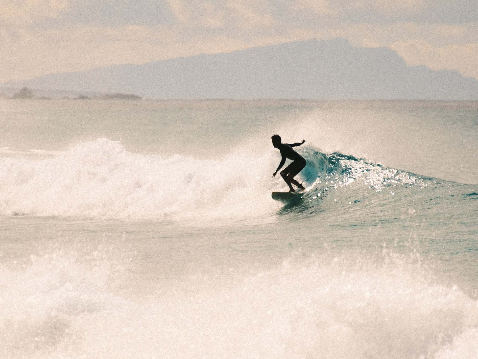 Surfista a contraluz girando entre la espuma, con la silueta de una isla al fondo