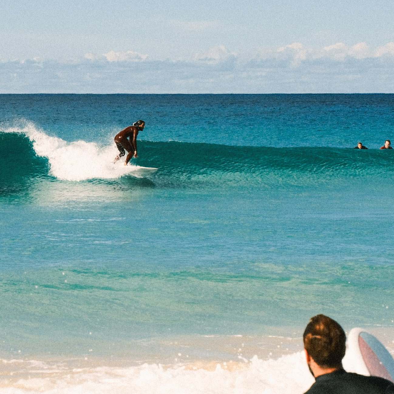 Surfer riding a turquoise wave, watched by a friend from the shore