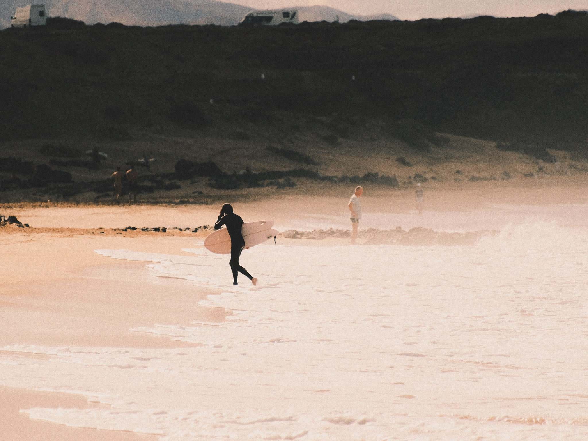 Surfista entrando al mar con la tabla en una playa de arena y roca volcánica
